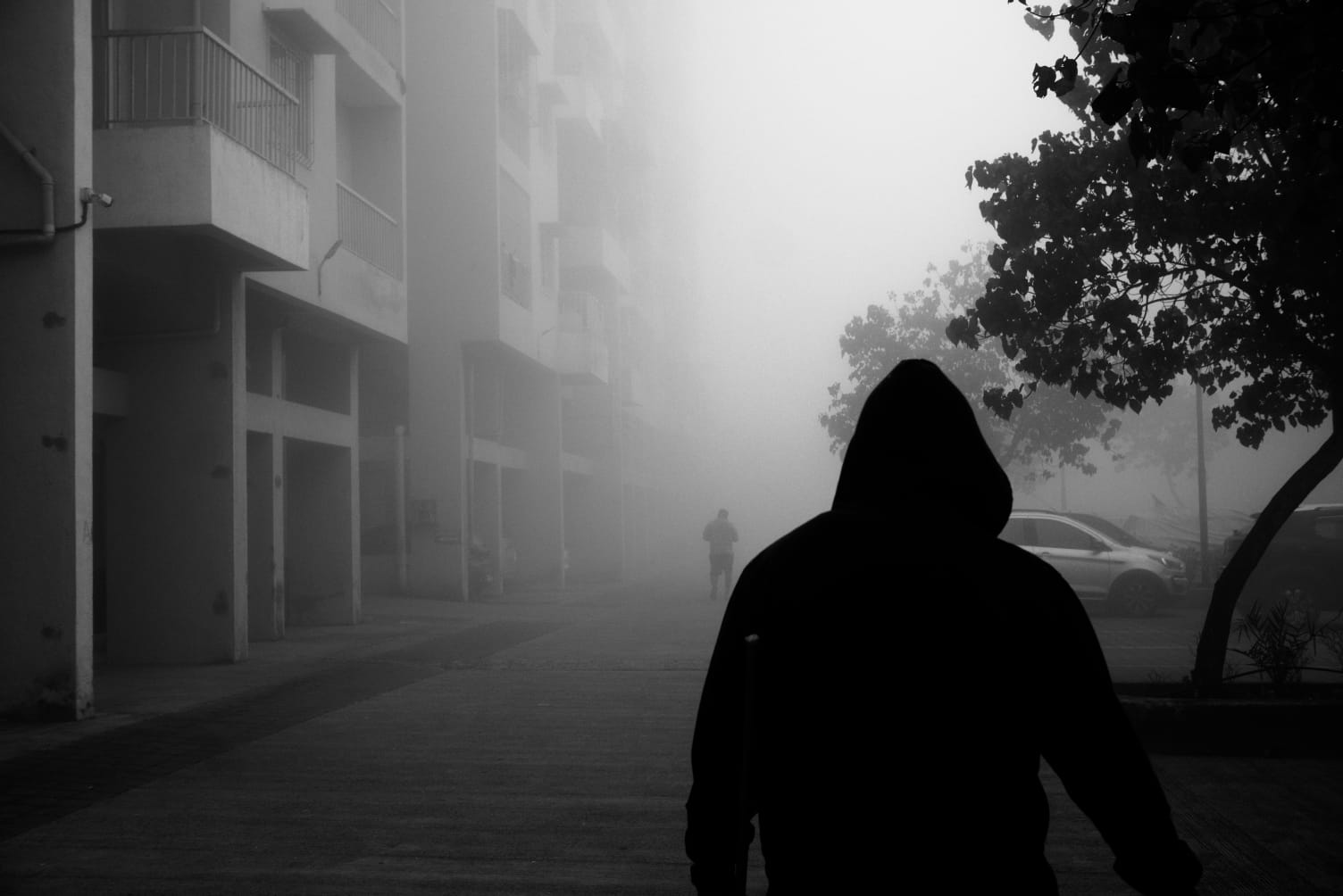 A hooded figure in silhouette walking into heavy fog, a second figure barely visible ahead