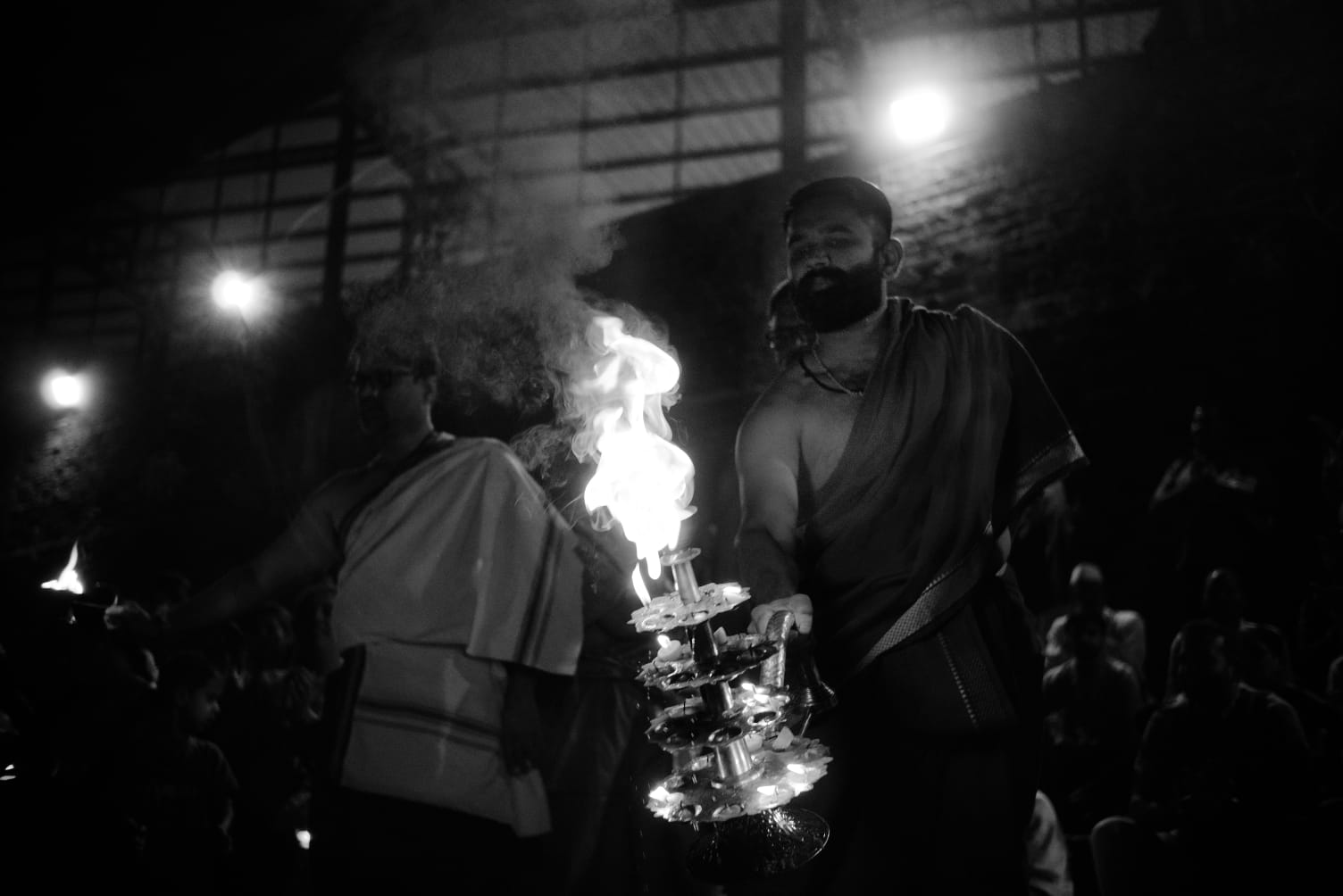 A man carrying a tiered oil lamp through a crowd at night, flame rising from the top