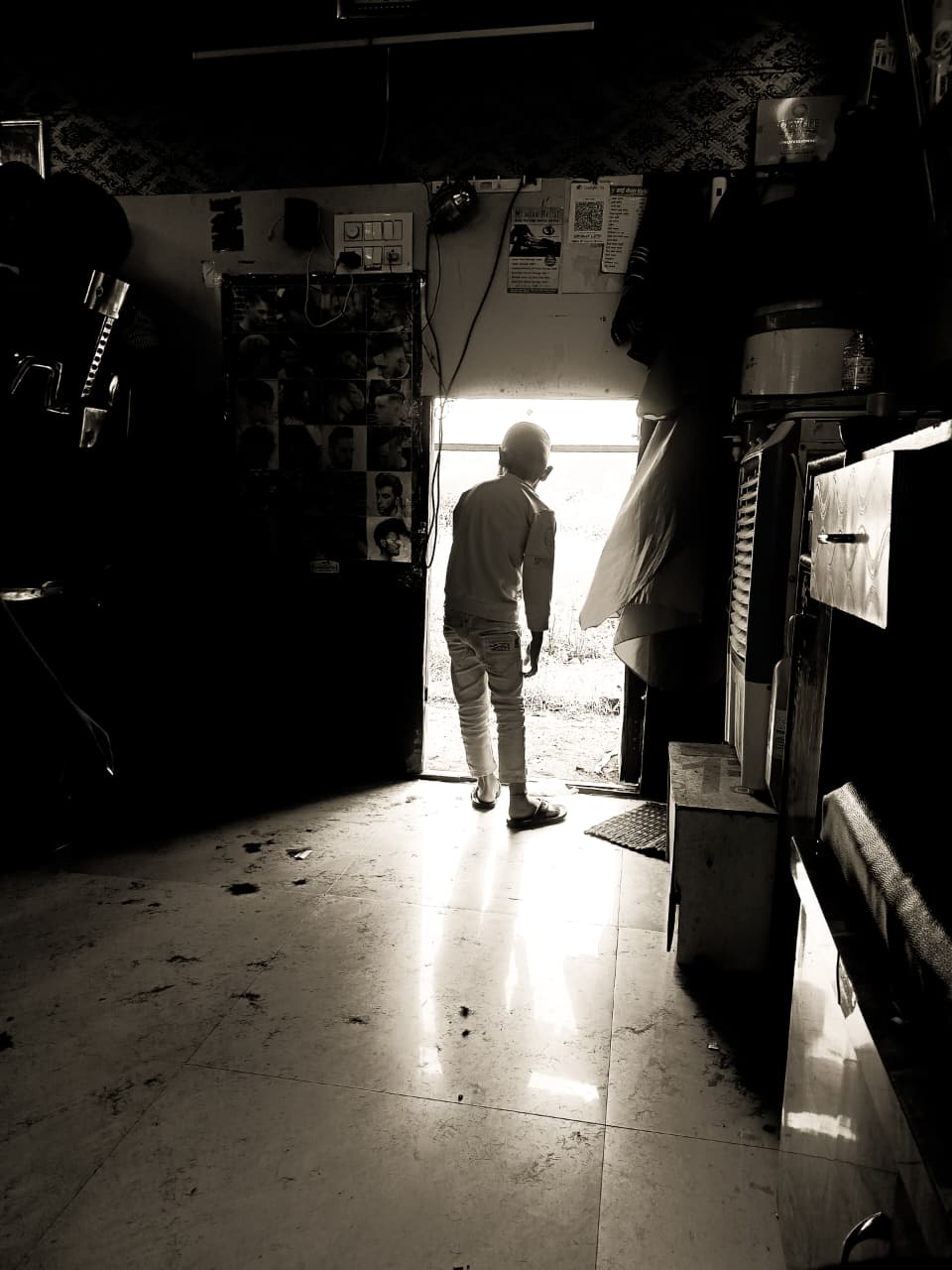 A boy standing in a barbershop doorway, backlit against bright daylight, haircut posters on the dark wall