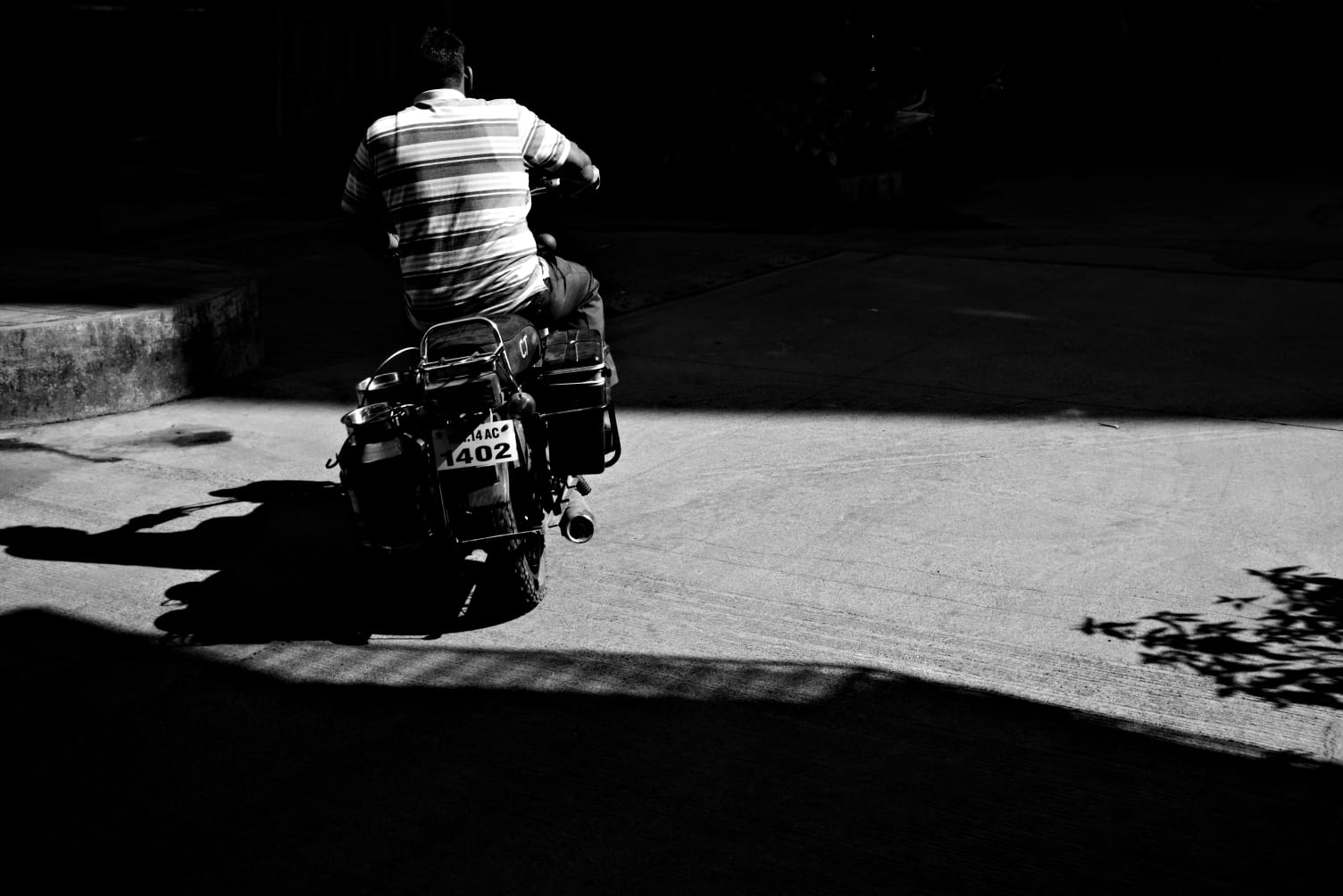 A man riding a motorcycle across a hard band of sunlight, his shadow thrown long behind him