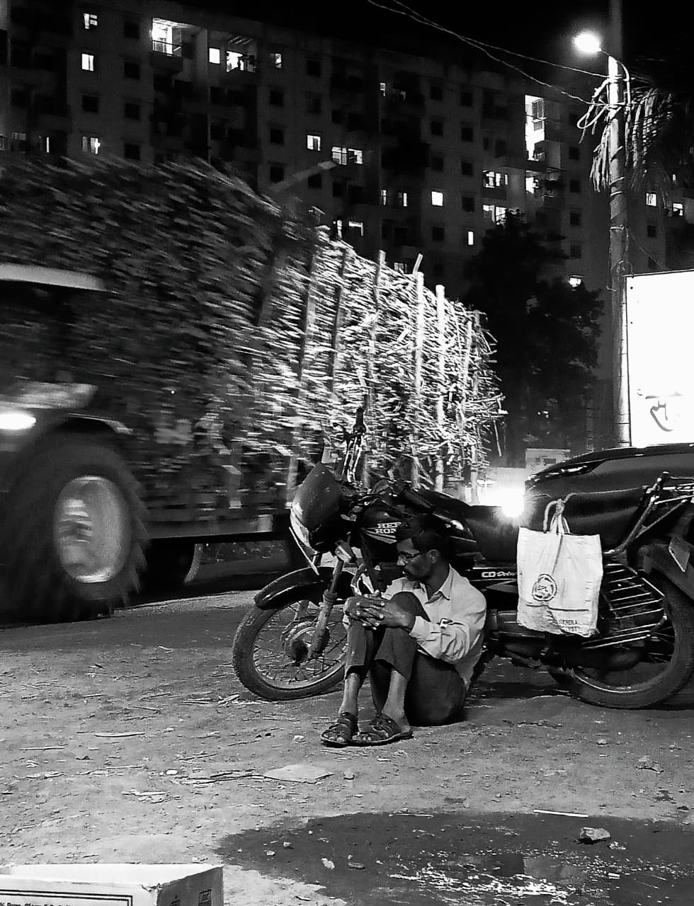 A man sitting on the ground beside a parked motorcycle as a sugarcane truck streaks past, lit tower block behind