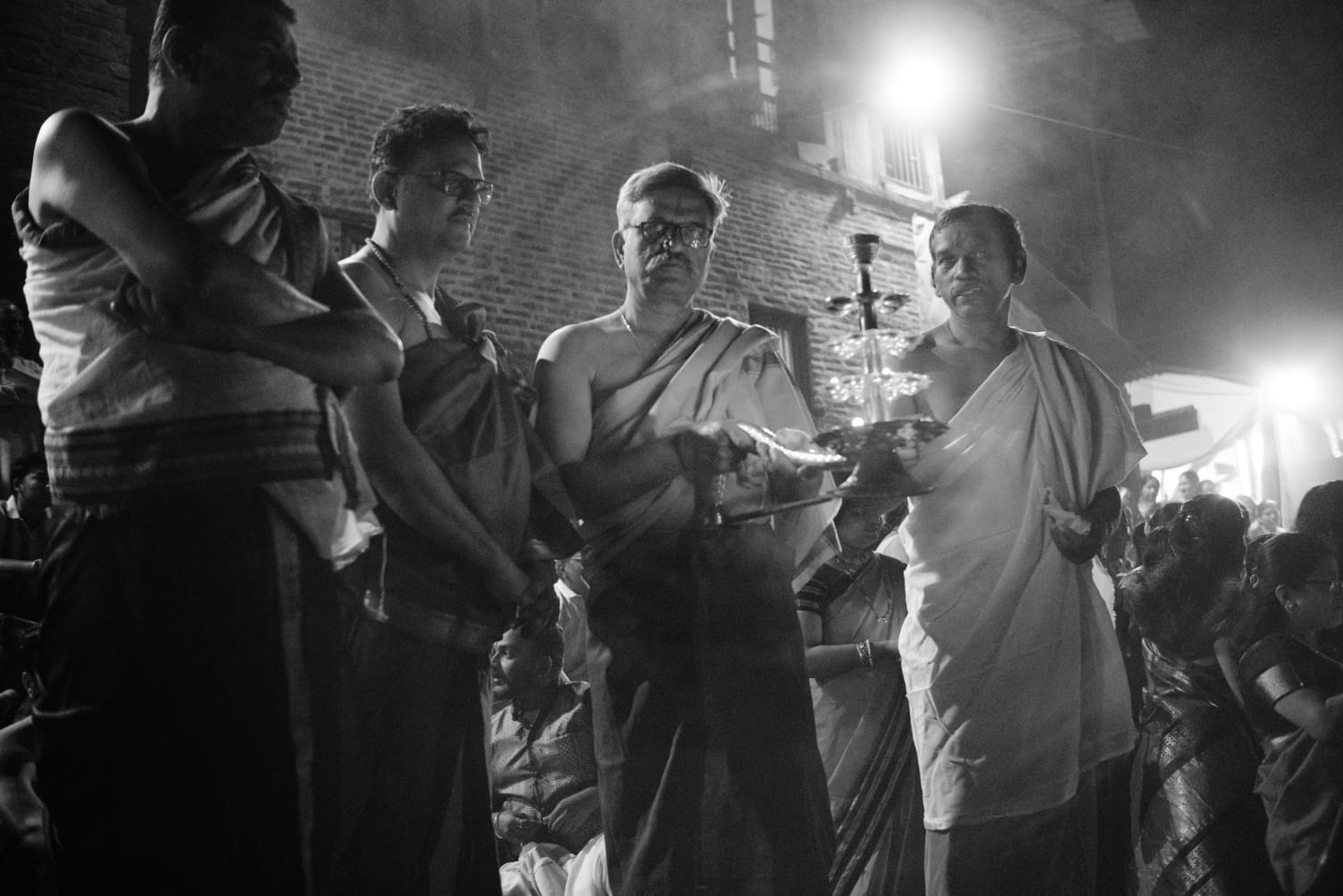 Priests holding lamps during an evening ritual, smoke drifting through the light